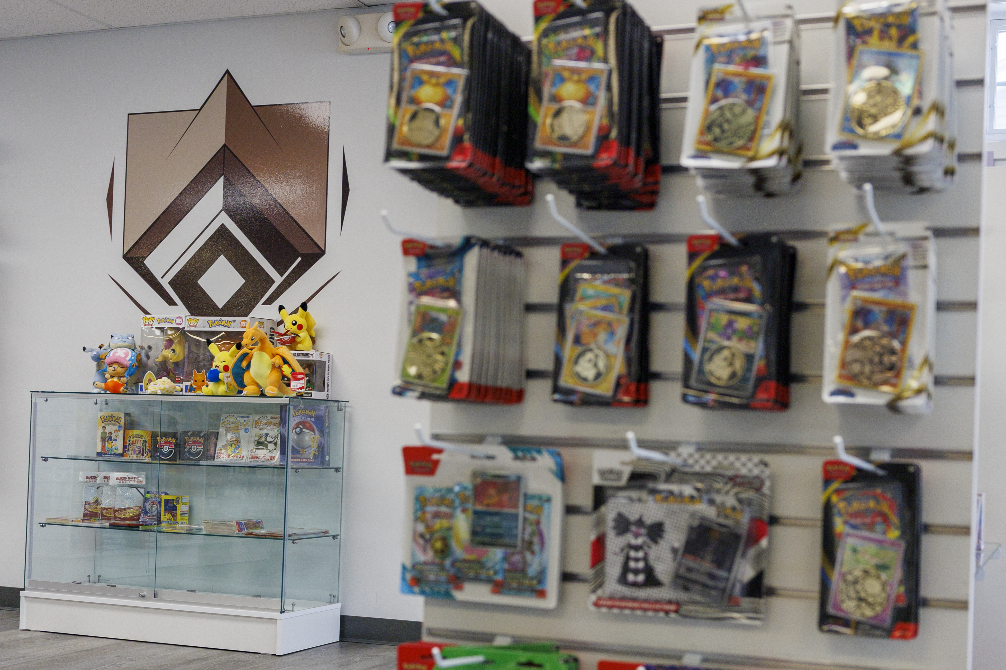Kadara Cards shop interior with the store logo on the wall, a glass display case of sealed products and plushies, and a wall of Pokémon blister packs.
